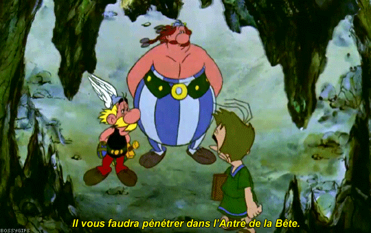 Capture d'écran tirée d'un dessin animé d'Astérix. La scène montre Astérix, Obélix et un petit guide en tunique verte debout dans une vaste grotte sombre aux parois rocheuses et verdâtres. Au centre, Obélix, grand et fort avec ses tresses rousses et son pantalon à rayures bleues et blanches, se tient les mains sur les hanches et regarde d'un air hébété vers le haut de la caverne. À sa gauche, le petit guerrier Astérix, portant sa moustache blonde et son casque ailé, écoute attentivement. À droite, le petit homme aux cheveux bruns s'adresse à eux, de profil, en tenant un dossier sous le bras. Au bas de l'image, un sous-titre jaune affiche le dialogue : « Il vous faudra pénétrer dans l'Antre de la Bête. »